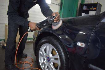 Auto polijstproces dichtbij opname met oranje elektrische polijstmachine en zwart voertuig bij wiel in garage omgeving