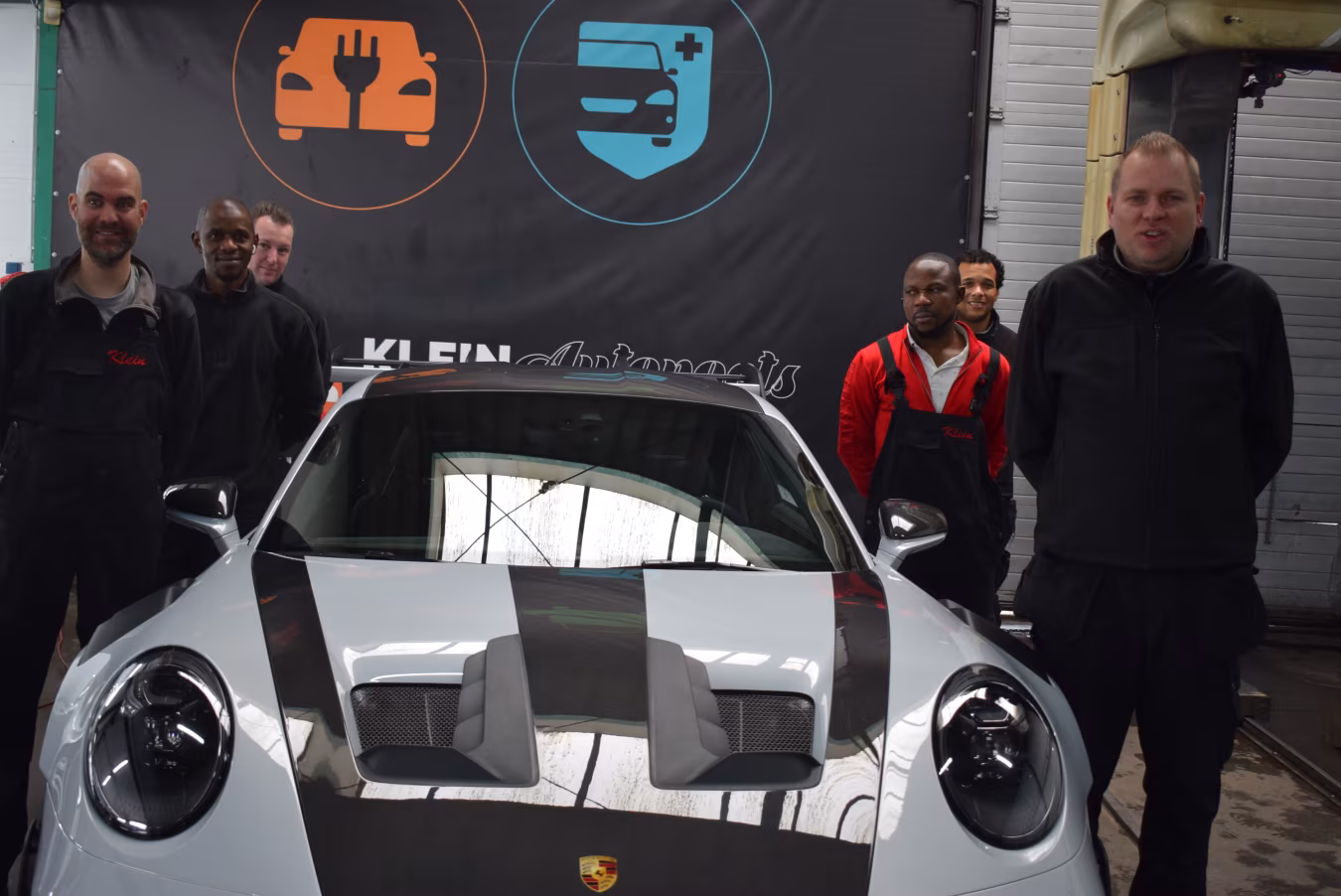 Ontmoet het team bij Klein Autopoets met een Porsche sportwagen en banner KLEIN Autosport in garage