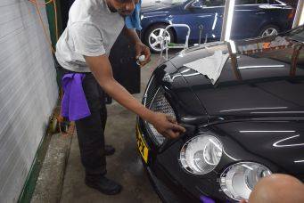 Interieur werkplaats met glanzende zwarte auto en detailwerk aan grille door medewerker, focus op autoreparatie en onderhoud