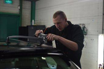 H2 titel autoverzorging: iemand gebruikt een elektrische polijstmachine op een autodak in een garage met groene deur en verlichting