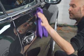 Binnenruimte met zwarte auto in werkplaats, hand met paarse doek poetst auto glanzend met reflectie gezicht, autopoets groningen