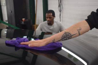 Tatoeage met kompas en buena op arm die paarse doek tegen glanzende autolak drukt tijdens poetsen in garage klein autopoets groningen