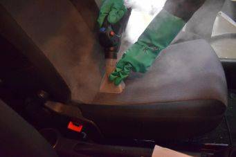 Close-up van een autostoel die gereinigd wordt met stoom, groene handschoenen en stoomreinigingsapparaat zichtbaar in actie